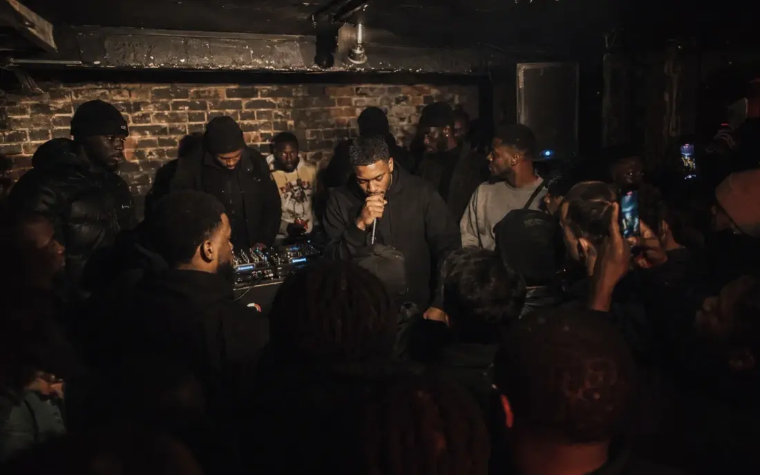 A rapper performing in a brick basement, phones and hands in the air.