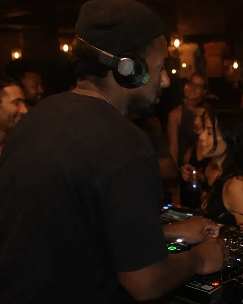 A DJ at the decks, headphones on, the dance floor moving behind.